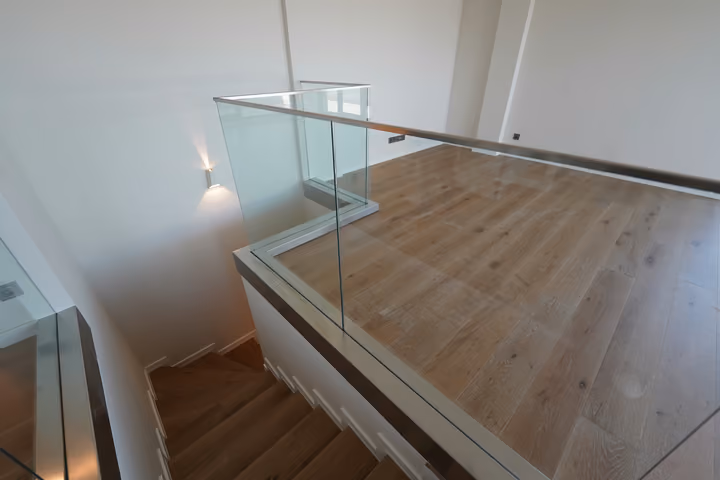 An internal wood-finished staircase and landing edged with a glass balustrade.