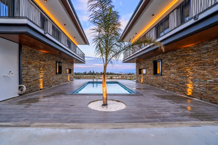 Evening view of the shared pool and central palm tree between two stone-clad villas, with balconies and warm exterior lighting.