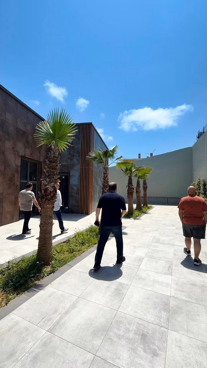 A paved walkway, palm planting and people beside a building. Undated photograph supplied in the construction source folder; it does not establish the current completion status.