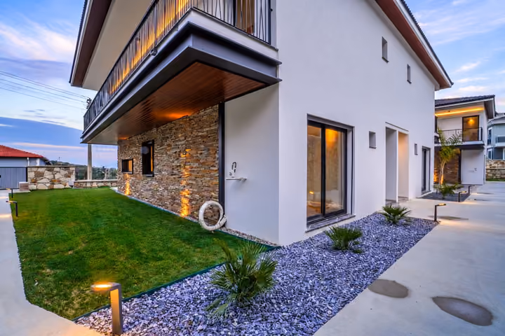 The villa corner shows an upper balcony, stone and white facades, garden doors, a lawn and a lit path with low plants.