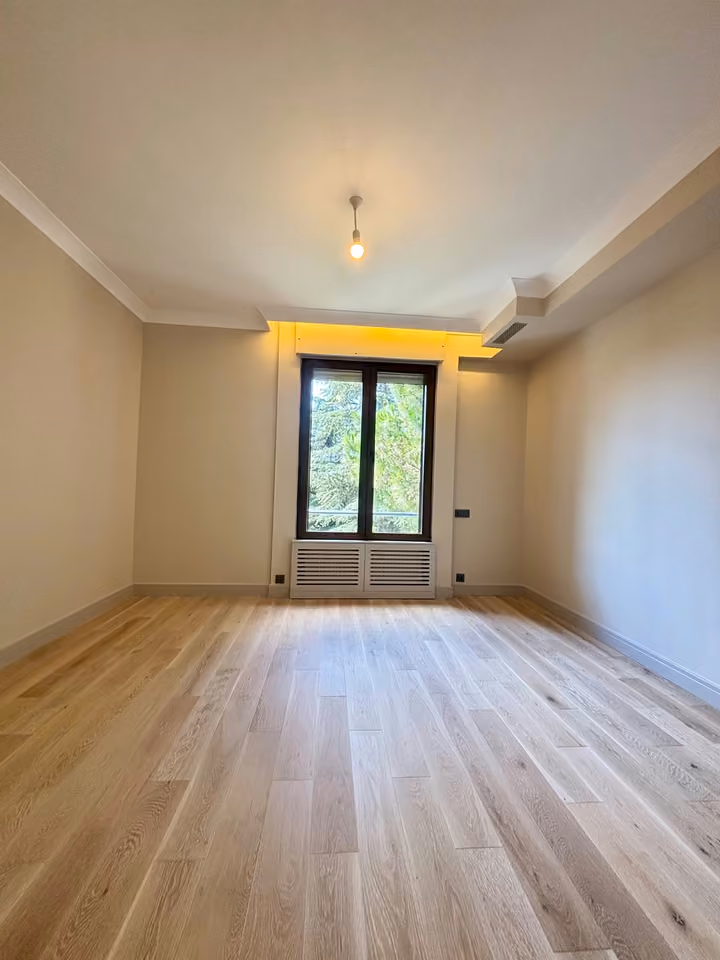 Full view of a bedroom with pale flooring and a tall window.