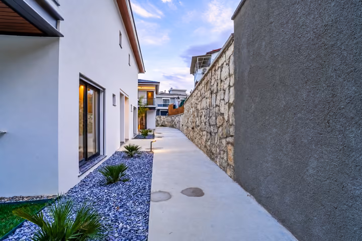 A paved side path runs between the white villa facade and a stone boundary wall, with low planting and garden lights.