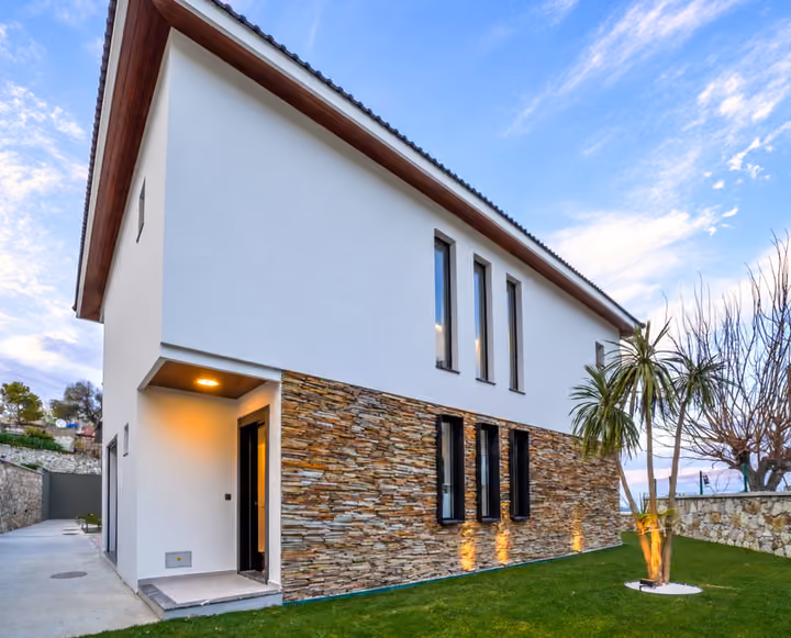 Two-storey villa exterior with a recessed entrance, stone cladding, narrow windows and a lawn with palms at dusk.