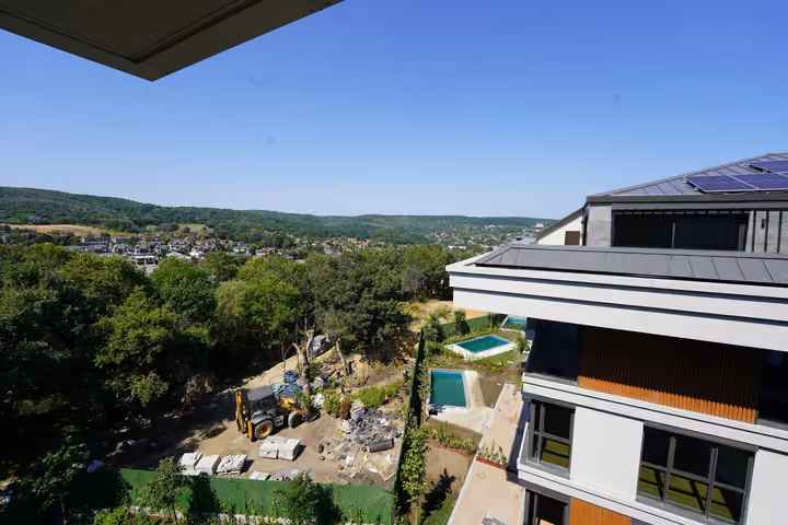 Trees and wooded hills seen from the apartment, with a neighboring facade and ground works in the foreground.