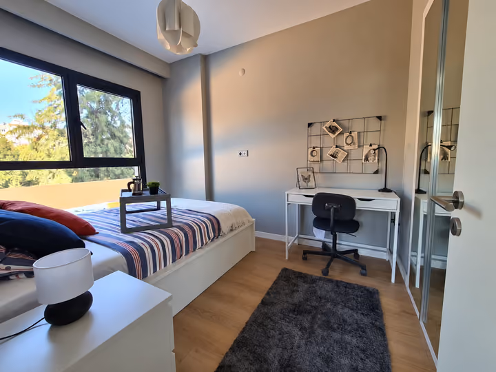 Photograph of a bedroom with a large window, bed and study desk.
