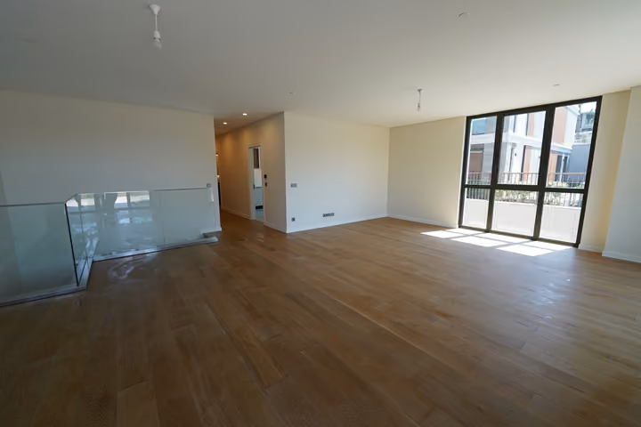 Photograph of an unfurnished living room with timber flooring, glazing and a glass stairwell balustrade in Zekeriyakoy.
