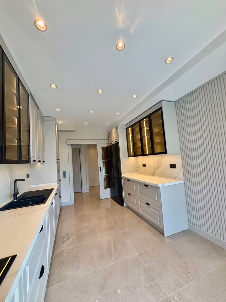Reverse view of the kitchen showing white cabinetry and illuminated glass-front cabinets.