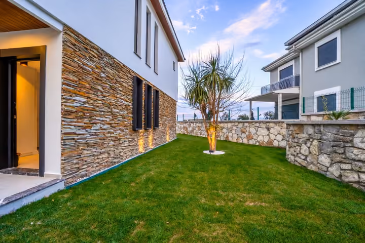 A lawn and planted tree beside the villa entrance, with stone cladding, tall windows and a low boundary wall.