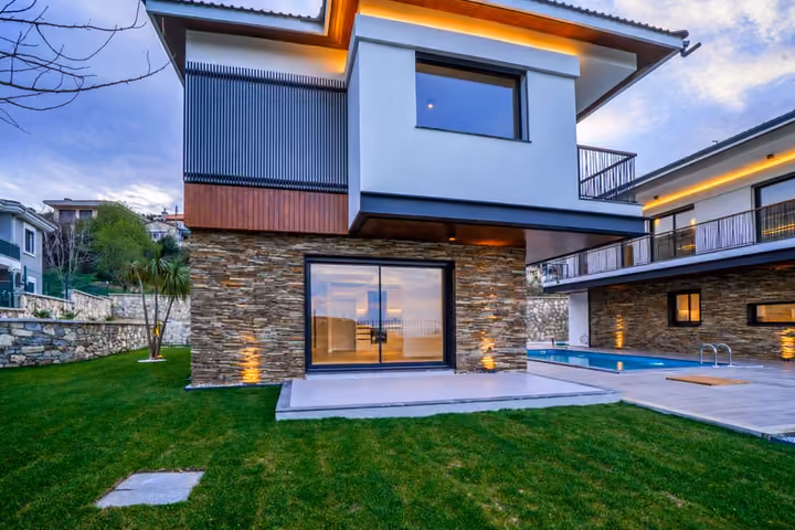 Garden view of the villa's glazed ground-floor opening, stone and white facade, lawn and adjacent shared pool.