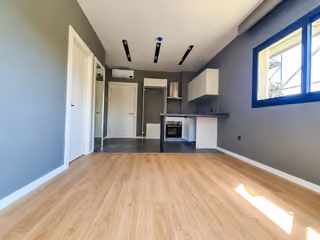 Photograph of an unfurnished room with an open kitchen, wood flooring and a window.