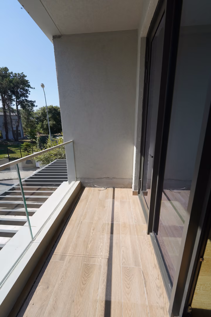 A narrow balcony with wood-effect floor tiles, glass railing and a glazed room entrance.