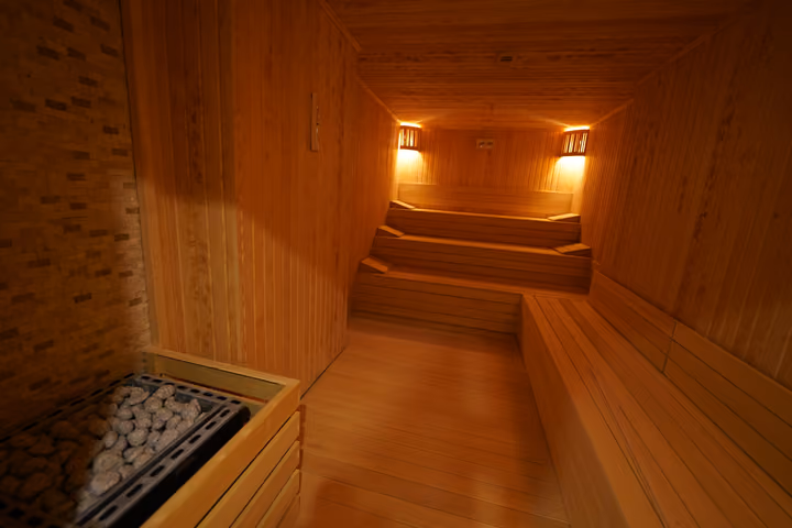 Interior view of a wood-lined sauna showing the heater, stepped seating and wall lights.