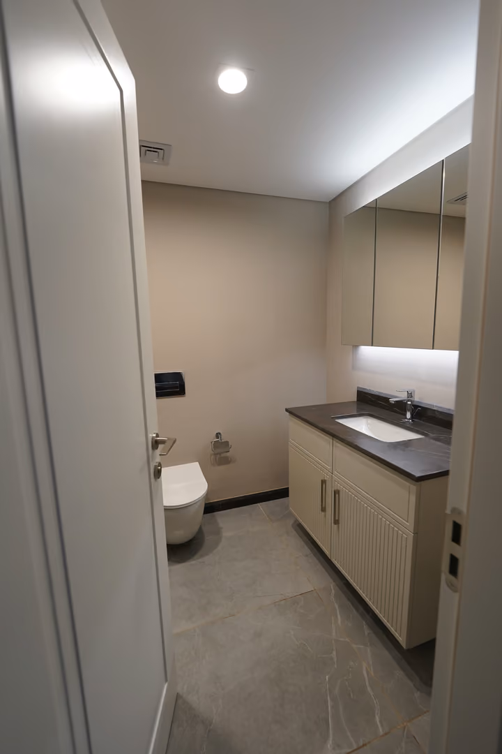 A second bathroom view showing a pale vanity, illuminated mirror and wall-mounted toilet.