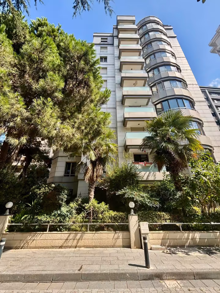 Apartment building facade with balconies and mature trees in front.