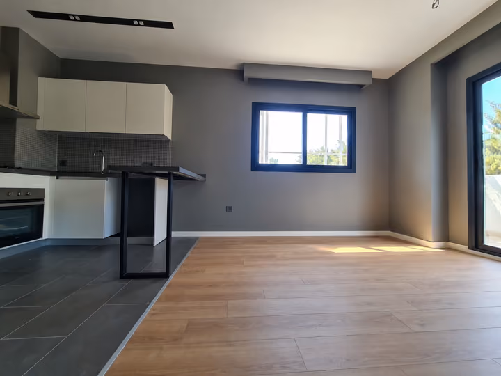 Photograph of an unfurnished room with wood flooring beside an open kitchen.