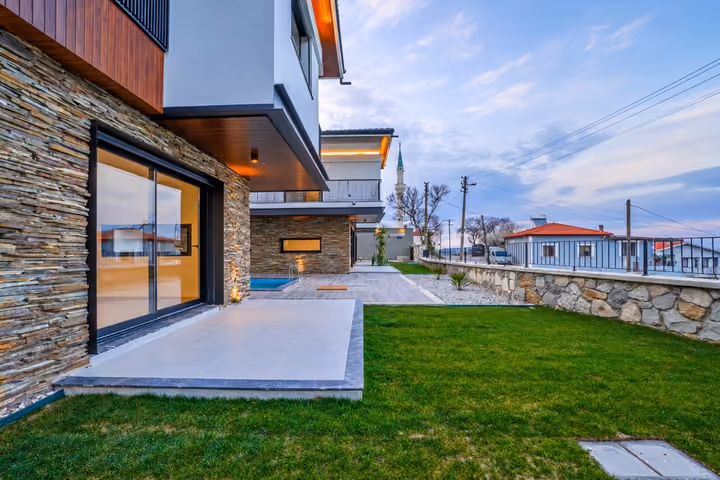 Lawn beside a stone-clad villa, a covered tiled terrace, sliding glass doors and a pool beyond the terrace.