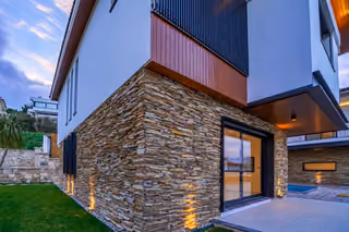 Stone-clad villa corner with a sliding glass door, tiled terrace, timber balcony panel and a pool visible to the side.