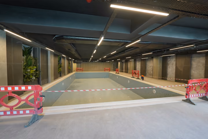 Wide view of an empty indoor pool, surrounding tiled deck and temporary safety barriers. Source photograph dated 22 July 2026.