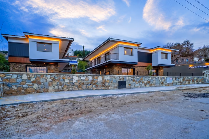 Wide street view of the two-storey villas, their balconies and illuminated roof edges behind a low stone boundary wall.
