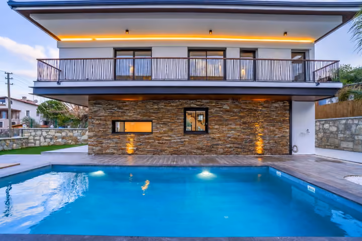 Frontal view of a villa with a full-width balcony, stone-clad lower facade and exterior lighting beside the blue swimming pool.